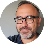 A circular headshot of a middle-aged man with short dark hair, a salt-and-pepper beard, and glasses. He is smiling slightly at the camera, wearing a navy blue shirt against a neutral grey background.