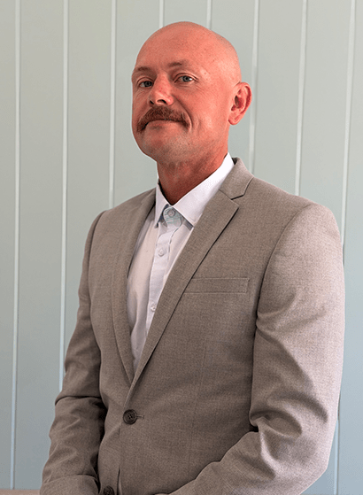 A professional portrait of Chris Cavanagh, founder of True Blue. He is a man with a shaved head and a mustache, dressed in a light grey blazer over a white button-down shirt, posing against a pale blue wood-paneled wall.