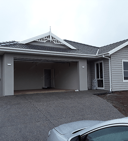 A modern single-story home with light grey horizontal siding and a dark tiled roof. The house features a decorative white gable finial, an open grey garage, and a pebble-crete driveway.