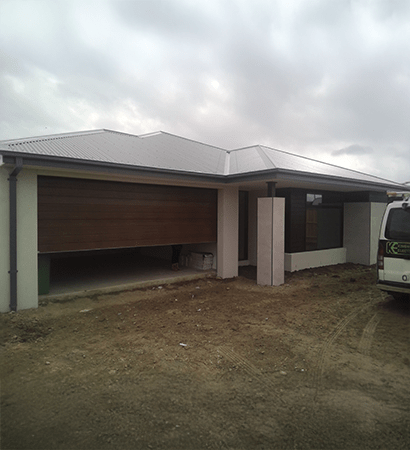A contemporary single-story home with white exterior walls and a light grey corrugated metal roof. The house features a wooden garage door partially opened and a large front window, set against a dirt yard under an overcast sky.