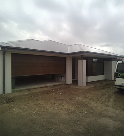 A contemporary single-story home with white exterior walls and a light grey corrugated metal roof. The house features a wooden garage door partially opened and a large front window, set against a dirt yard under an overcast sky.