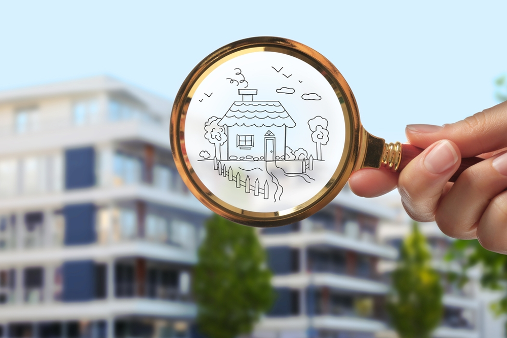 A hand holds a gold-rimmed magnifying glass in front of a blurred modern apartment building. Inside the magnifying glass is a simple line drawing of a cozy cottage with a chimney, fence, and trees, symbolizing the search for a dream home.