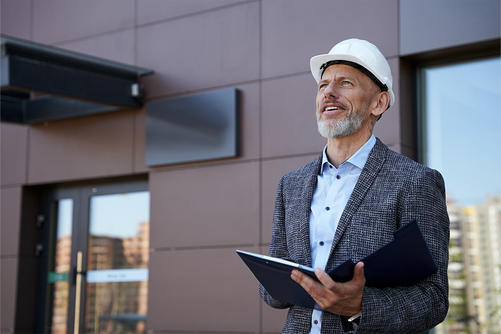 A professional building inspector with a grey beard, wearing a white hard hat and a grey blazer over a blue shirt. He is holding an open blue folder and looking upward at a modern building facade with a satisfied or thoughtful expression.