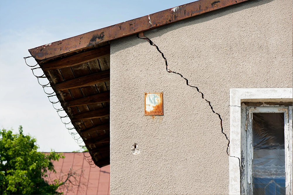 A close-up of a significant diagonal structural crack running down the exterior stucco wall of an older building. The damage starts from the roofline near a rusted metal gutter and extends toward a window frame, illustrating a serious maintenance issue or foundation settling.