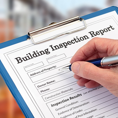 A close-up shot of a person’s hand holding a silver pen, about to fill out a "Building Inspection Report" form. The document is secured on a blue clipboard and includes sections for property address, owner name, and inspection results such as general condition and evidence of leaks.