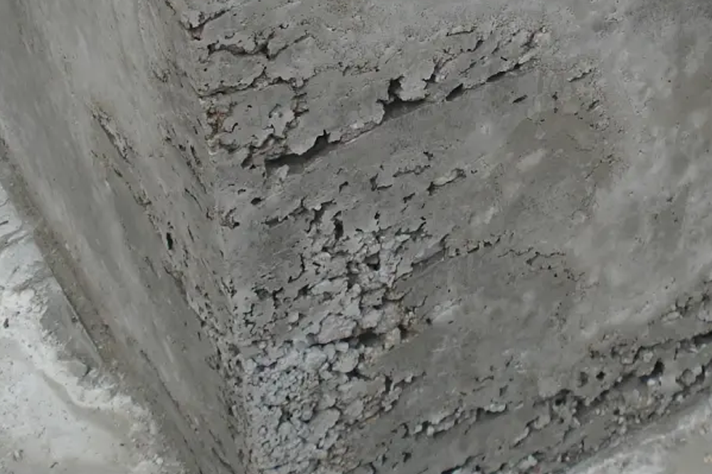 A close-up of a concrete pillar showing significant "honeycombing" defects. The surface is rough and pitted with deep air pockets and exposed aggregate, indicating poor compaction or a faulty pour.