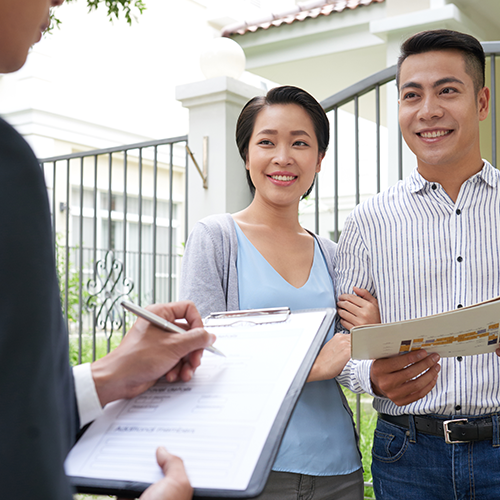 A professional in a suit holds a clipboard and pen, appearing to go through a checklist with a smiling young couple standing in front of a residential property.