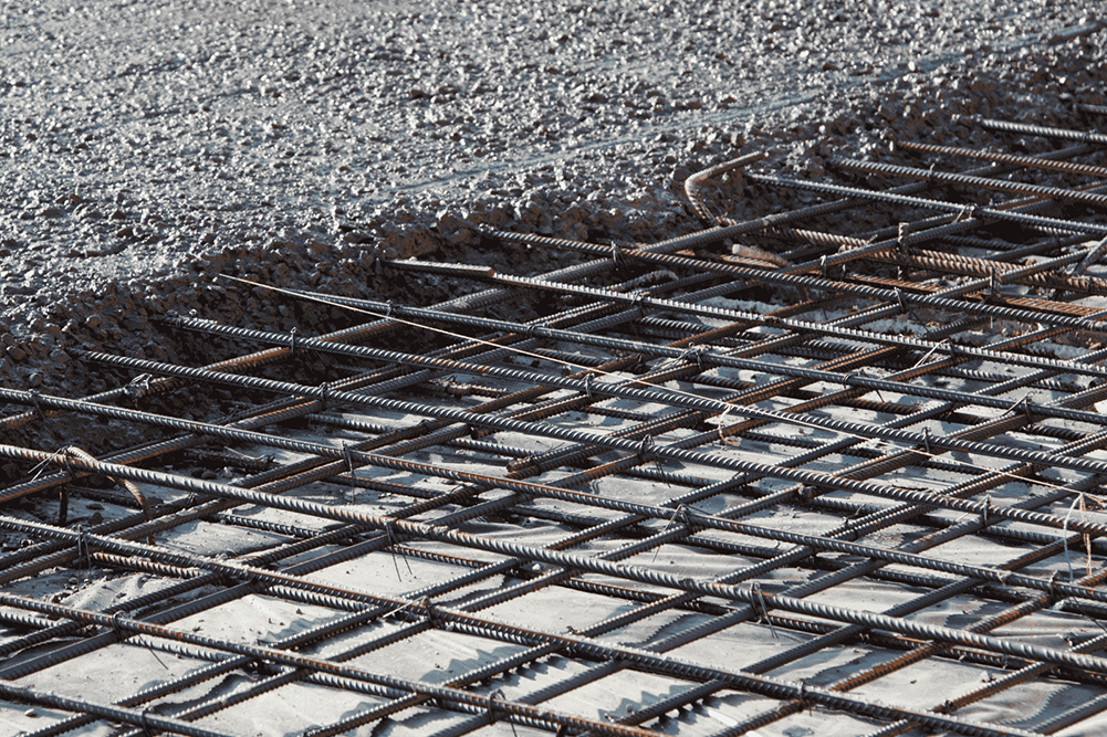 A close-up of a concrete slab being poured on a construction site. The image shows a section of wet, textured concrete meeting a grid of steel reinforcement rebar laid over a grey vapor barrier, highlighting the structural layering of a building foundation.