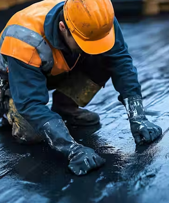 A construction worker in a bright orange hard hat, safety vest, and heavy-duty black gloves kneeling on a flat surface. He is manually smoothing down or inspecting a layer of wet, black liquid waterproofing membrane.