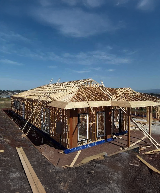 A wide-angle shot of a new home's timber frame under construction against a clear blue sky. The wooden wall studs and roof trusses are fully assembled, with window frames installed and blue moisture barrier wrap visible along the base of the structure.