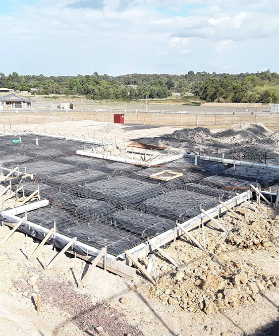 A wide, high-angle shot of a residential concrete slab foundation under preparation. The site features black vapor barrier plastic laid over the ground, covered by a grid of steel reinforcement mesh (rebar), all contained within wooden formwork. The background shows a large, open construction site under a clear sky.